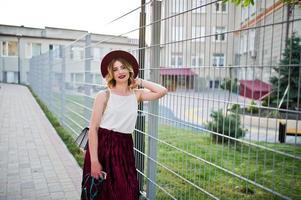 ragazza modello bionda alla moda e bella in elegante gonna in velluto di velluto rosso, camicetta bianca e cappello, in posa con telefono e auricolari. foto