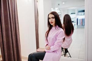 bruna splendida ragazza nella boutique del negozio di abbigliamento in abiti casual e camicetta rosa contro lo specchio. foto