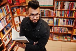 studente arabo alto e intelligente, indossa una giacca di jeans nera e occhiali da vista, in biblioteca con un libro a portata di mano. foto