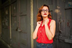 attraente donna dai capelli rossi in occhiali, indossare su camicetta rossa e gonna di jeans in posa. foto