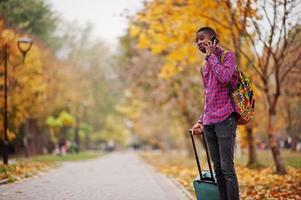 uomo afroamericano in camicia a scacchi, con valigia e zaino. viaggiatore dell'uomo di colore al parco d'autunno che parla sul telefono cellulare. foto