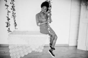 attraente donna afroamericana con capelli afro su gonna e giacca di jeans, posata in una stanza bianca sull'altalena. modello nero alla moda. foto