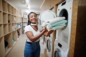 donna afroamericana allegra con asciugamani in mano vicino alla lavatrice nella lavanderia self-service. foto