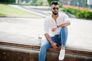 elegante uomo arabo alto modello in camicia bianca, jeans e occhiali da sole seduto in strada della città. barba attraente ragazzo arabo con una tazza di caffè al parco. foto