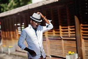 elegante uomo afroamericano in camicia bianca e pantaloni colorati con cappello e occhiali in posa all'aperto. ragazzo nero modello alla moda. foto