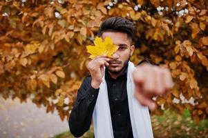 uomo indiano alla moda in abiti tradizionali neri con sciarpa bianca in posa all'aperto contro l'autunno giallo e mostra il dito. foto