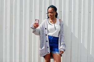 donna africana con i capelli dreadlocks, in pantaloncini di jeans posati contro il muro di acciaio bianco e facendo selfie per telefono. foto