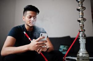 uomo indiano bello e alla moda in nero seduto in camera, fumando narghilè e guardando sul cellulare. foto