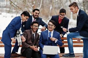 un gruppo di sei uomini d'affari indiani in giacca e cravatta poste all'aperto in una giornata invernale in Europa, guardando sul laptop e ridendo.