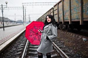 ragazza bruna in cappotto grigio con ombrello rosso nella stazione ferroviaria. foto