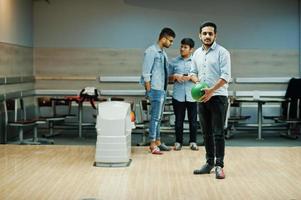 uomo del sud asiatico in camicia di jeans in piedi alla pista da bowling con la palla sulle mani. ragazzo si sta preparando per un tiro. gli amici lo supportano ad alta voce. foto