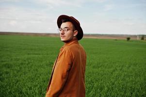 uomo alla moda con gli occhiali, giacca marrone e cappello in posa sul campo verde. foto