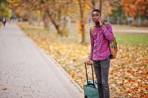 uomo afroamericano in camicia a scacchi, con valigia e zaino. viaggiatore dell'uomo di colore al parco d'autunno che parla sul telefono cellulare. foto