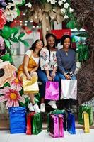 belle tre ragazze afro americane ben vestite con borse della spesa colorate sedute su una zona fotografica di decorazione primaverile nel centro commerciale. foto