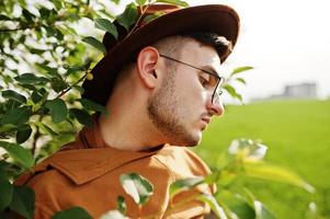 uomo alla moda con gli occhiali, giacca marrone e cappello in posa sul campo verde. foto