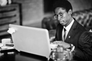 uomo d'affari afroamericano indossa su abito nero e occhiali seduto in ufficio con laptop e lavoro. foto