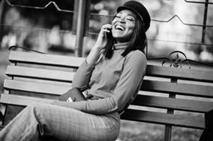 ragazza di moda afroamericana in berretto da strillone, con borsetta in posa in strada, seduta su una panchina con il telefono cellulare. foto