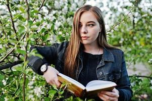 giovane ragazza bruna in jeans contro albero in fiore di primavera leggere il libro. foto
