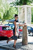 ragazza afroamericana in occhiali da sole, abito bianco e nero e borsa in posa contro il parcheggio delle auto. foto
