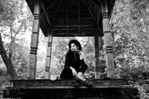 ragazza sensuale tutta in nero, labbra rosse e cappello. goth drammatica donna seduta su arco di legno nella foresta. ritratto in bianco e nero. foto