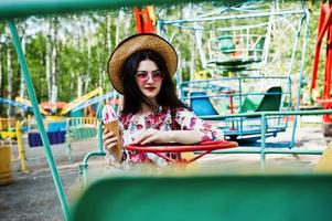 ritratto di ragazza bruna con occhiali rosa e cappello con gelato al parco divertimenti. foto