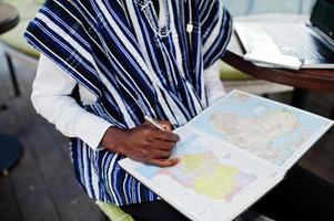 uomo africano in abiti tradizionali seduto dietro il laptop al caffè all'aperto e guardando la mappa dell'africa e del ghana sul suo taccuino. foto