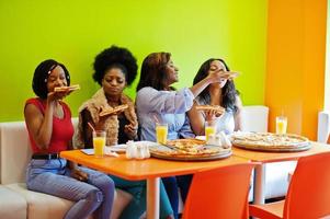 quattro giovani ragazze africane in un ristorante dai colori vivaci che mangiano fette di pizza in mano. foto