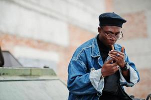 uomo afroamericano in giacca di jeans, berretto e occhiali, accende un sigaro e posa contro un veicolo blindato militare btr. foto