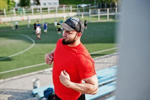 giovane uomo muscoloso barbuto brutale indossa maglietta rossa, pantaloncini e berretto allo stadio. foto