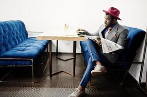 elegante uomo afroamericano modello in giacca grigia cravatta e cappello rosso bere caffè al bar e leggere i giornali. foto