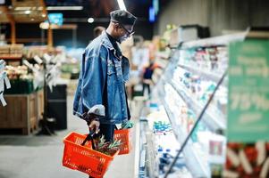 elegante uomo afroamericano casual con giacca di jeans e berretto nero con due cesti, in piedi vicino al frigorifero e shopping al supermercato. foto