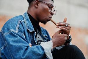 uomo afroamericano in giacca di jeans, berretto e occhiali, accende un sigaro. foto