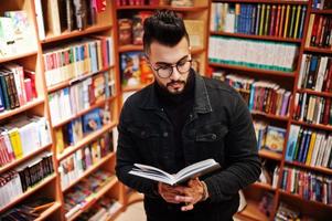 studente arabo alto e intelligente, indossa una giacca di jeans nera e occhiali da vista, in biblioteca con un libro a portata di mano. foto