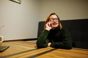 allegra giovane bella donna dai capelli rossi con gli occhiali, indossa un maglione di lana caldo verde, seduto al caffè. foto