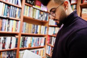 studente arabo alto e intelligente, indossa un dolcevita viola e occhiali da vista, in biblioteca leggi un libro. foto