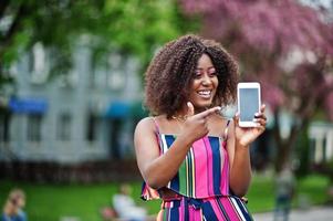 donna afroamericana alla moda in tuta a righe rosa posata in strada in fiore primaverile e mostra il dito sullo schermo del telefono cellulare. foto