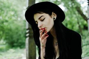 sensuale fumatrice tutta in nero, labbra rosse e cappello. donna drammatica goth che fuma sigaretta sottile. foto