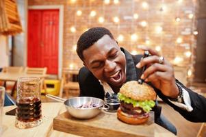 rispettabile giovane afroamericano in abito nero seduto al ristorante con gustoso doppio hamburger e bibita gassata. vuole mordere un pezzo. foto