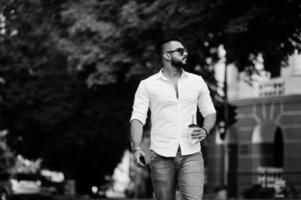elegante uomo arabo alto modello in camicia bianca, jeans e occhiali da sole posato in una strada della città. barba attraente ragazzo arabo con una tazza di caffè a piedi. foto
