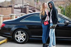 bella coppia multirazziale abbraccia insieme contro un'elegante auto sportiva nera. foto