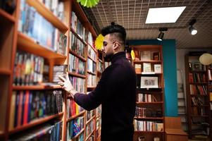 alto e intelligente studente arabo, indossa un dolcevita viola e occhiali da vista, in biblioteca che seleziona un libro sugli scaffali. foto