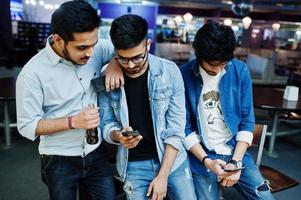 tre uomini asiatici alla moda indossano jeans guardando i telefoni cellulari e bevono bibite contro il tavolo del club. foto