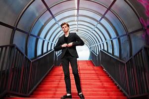 giovane ragazzo macho alla moda in giacca nera posato all'aperto di strada. incredibile modello uomo al tunnel delle scale rosse. foto