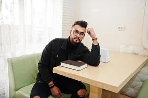 l'uomo arabo indossa una giacca di jeans nera e occhiali da vista seduti al bar, leggi il libro. ragazzo modello arabo elegante e alla moda. foto