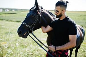 uomo arabo con barba alta indossa in nero e occhiali da sole con cavallo arabo. foto