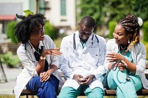 tre medici di gruppo afroamericani con uno stetoscopio che indossano un camice da laboratorio seduti su una panchina. foto