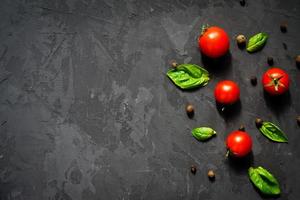succosi pomodorini maturi con foglie di basilico verde e pepe nero su sfondo nero. concetto di cibo salutare. vista dall'alto, copia spazio