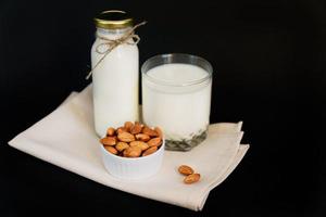 latte di mandorla fatto in casa in una bottiglia e bicchiere con noci in una ciotola di porcellana bianca su sfondo nero. foto