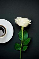 tazza di caffè nero e rosa bianca su sfondo nero, vista dall'alto, copia spazio. foto