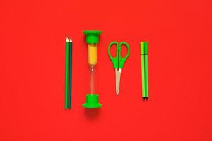 disposizione piatta creativa di materiale scolastico - penne verdi, matite, forbici e clessidra su sfondo rosso foto
